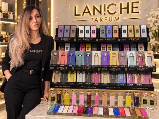 Laniche owner wearing black colour dress stands beside a large display rack filled with colorful perfume bottles and boxes under a lit LANCHE PARFÜM sign.
